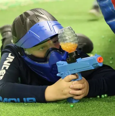 Un enfant masqué, allongé sur le terrain, vise avec son lanceur de billes de gel